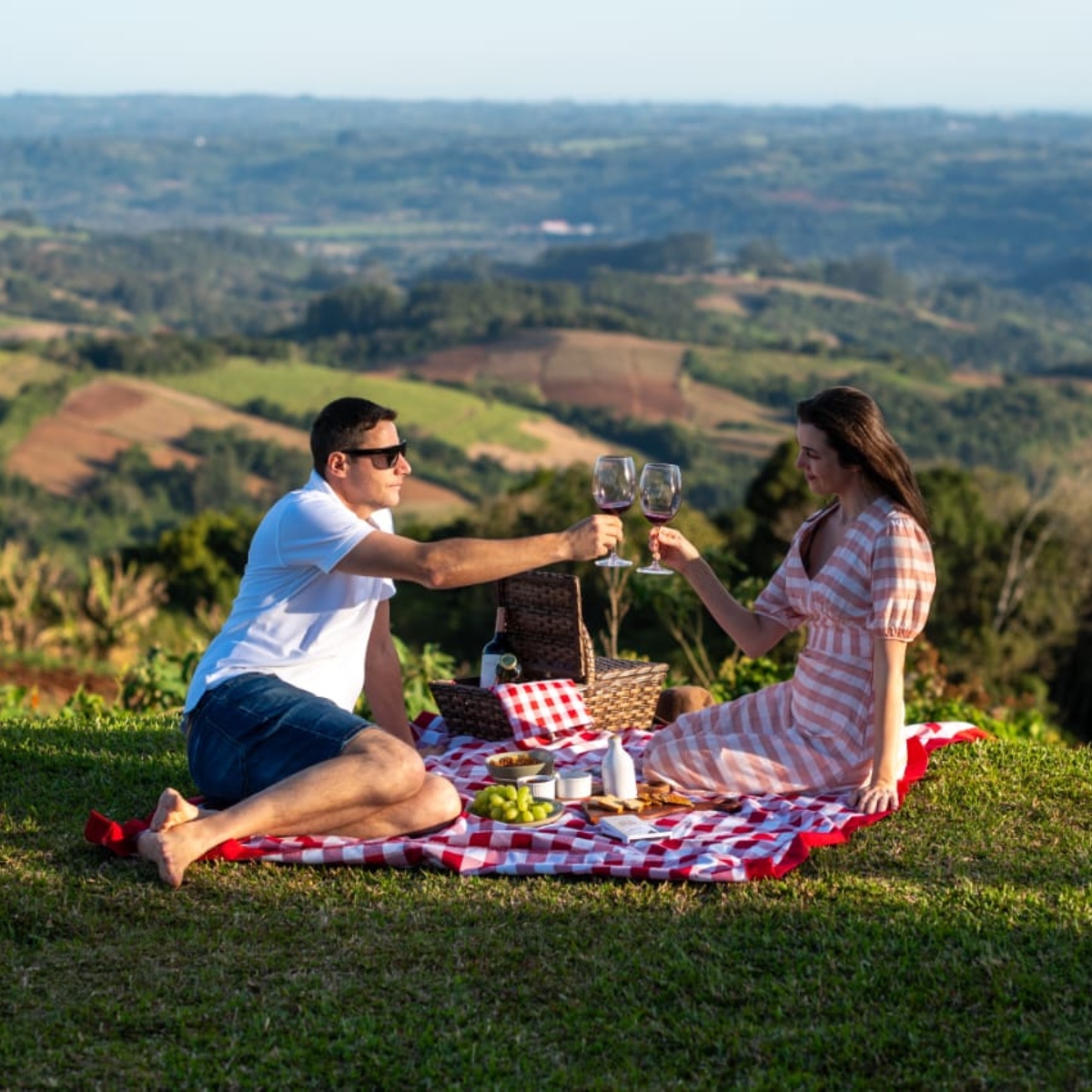 Casal em piquenique brindando com vista panorâmica das colinas gaúchas