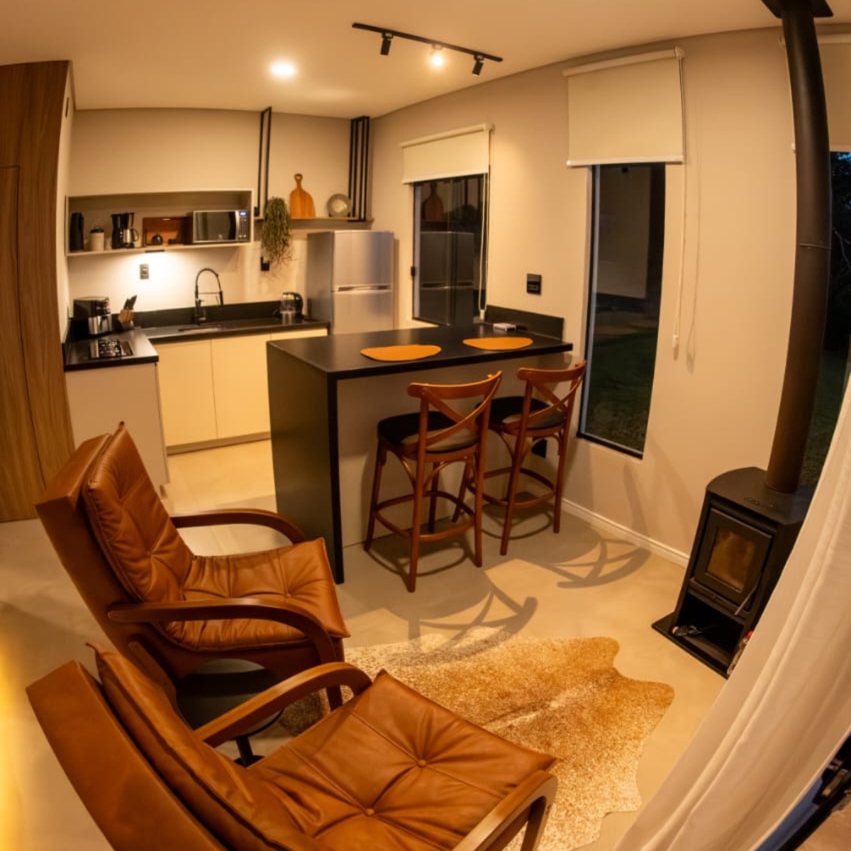 Sala de estar com poltronas de couro, calefator a lenha e cozinha equipada integrada