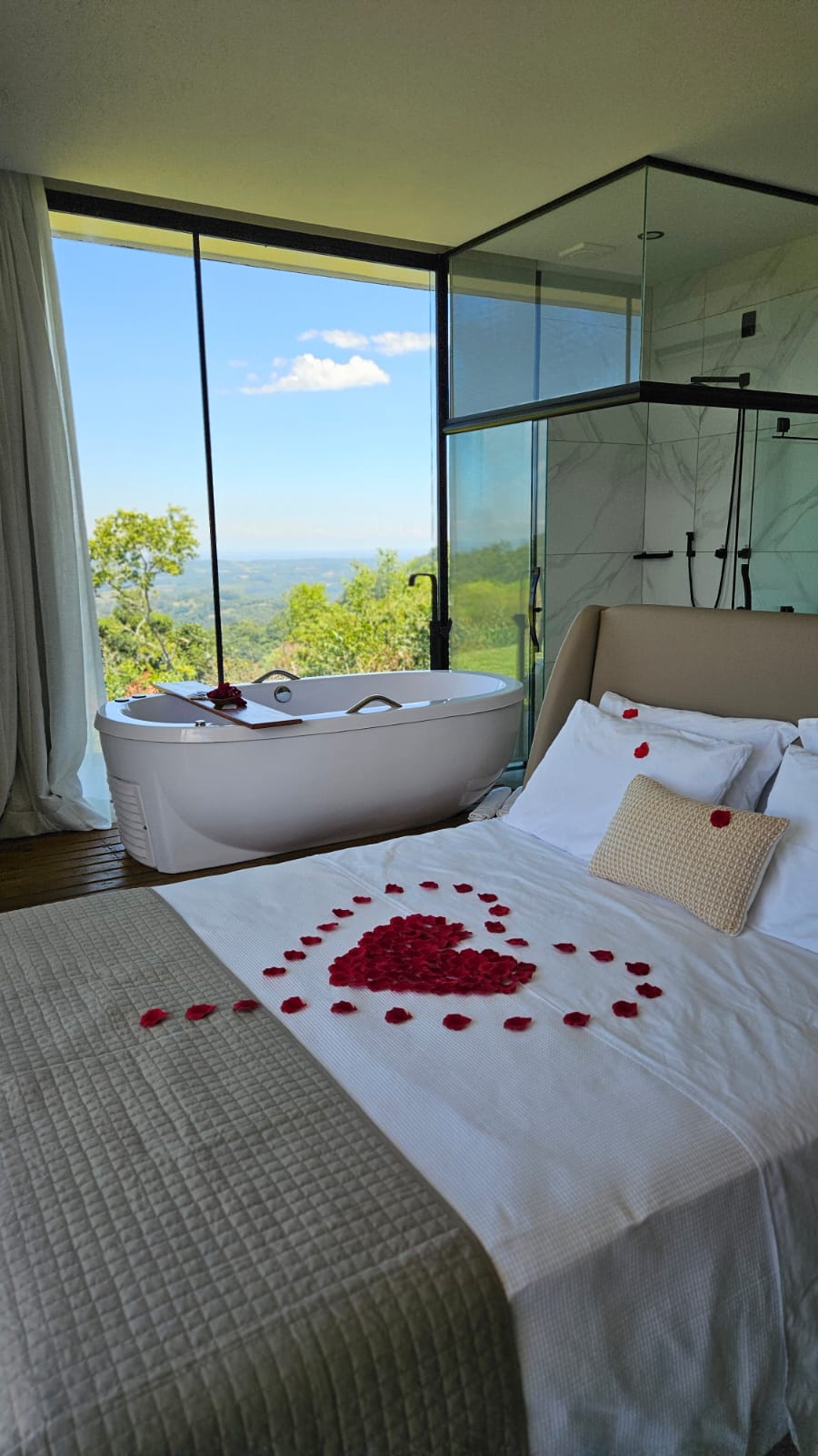Cama queen com decoração de pétalas de rosa e banheira com vista para a colina